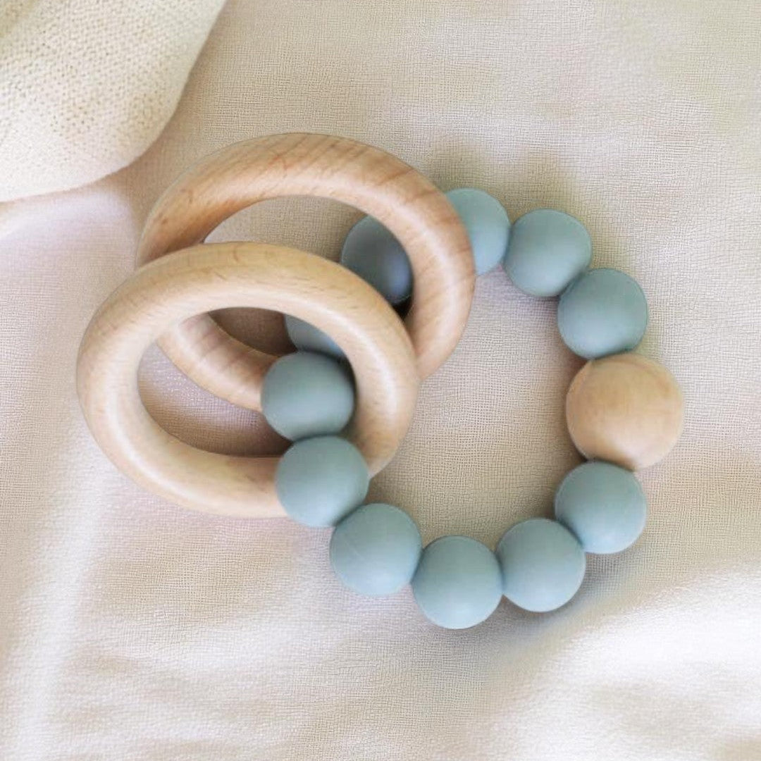 A set of two round beechwood and silicone teether rings in soft neutral colours, shown with a white cotton bag on a pale background.