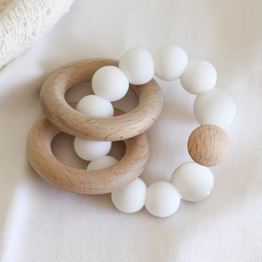 A set of two round beechwood and silicone teether rings in soft neutral colours, shown with a white cotton bag on a pale background.