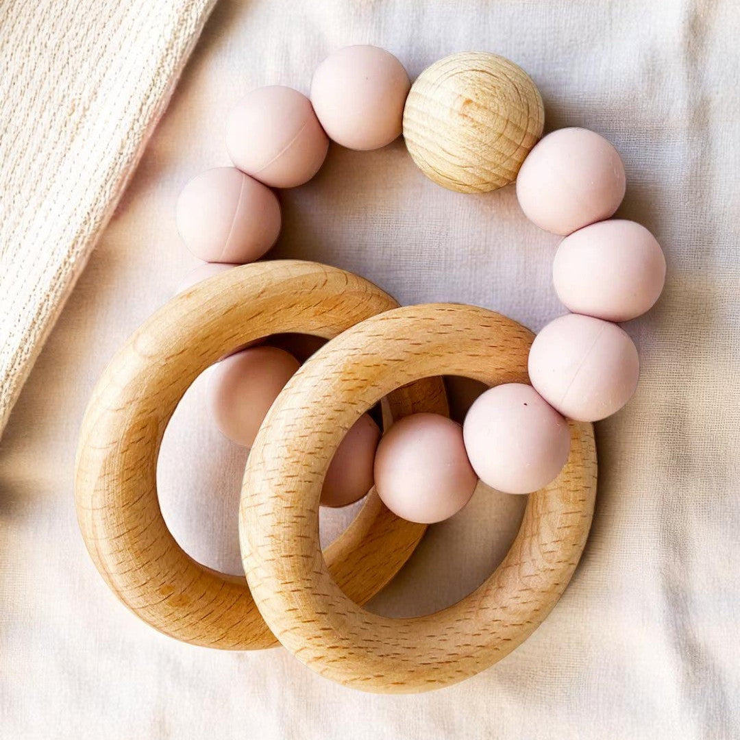 A set of two round beechwood and silicone teether rings in soft neutral colours, shown with a white cotton bag on a pale background.