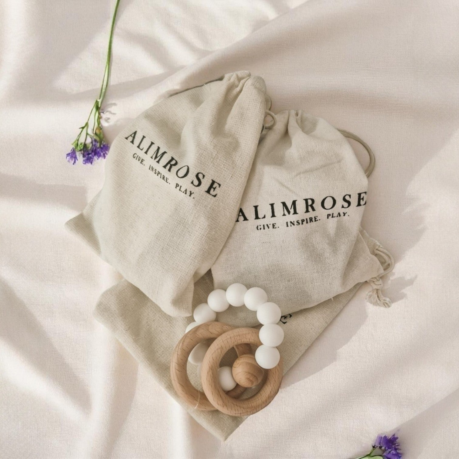 A set of two round beechwood and silicone teether rings in soft neutral colours, shown with a white cotton bag on a pale background.