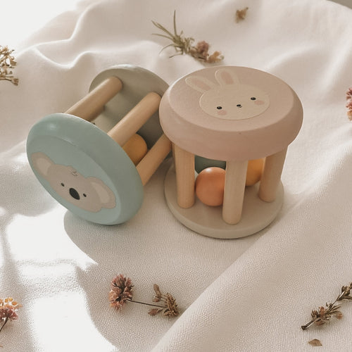 Two wooden baby rattles featuring pastel bunny and koala faces, with rolling beads inside, resting on a soft cream fabric with dried florals.